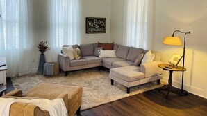 A living room with a sectional sofa, a small round table with a lamp, a ceiling fan, and a sign on the wall.