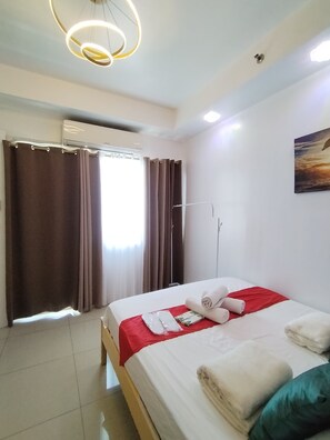 Una habitación de hotel con cama, cortinas, unidad de aire acondicionado, un cuadro en la pared y una lámpara en el techo.