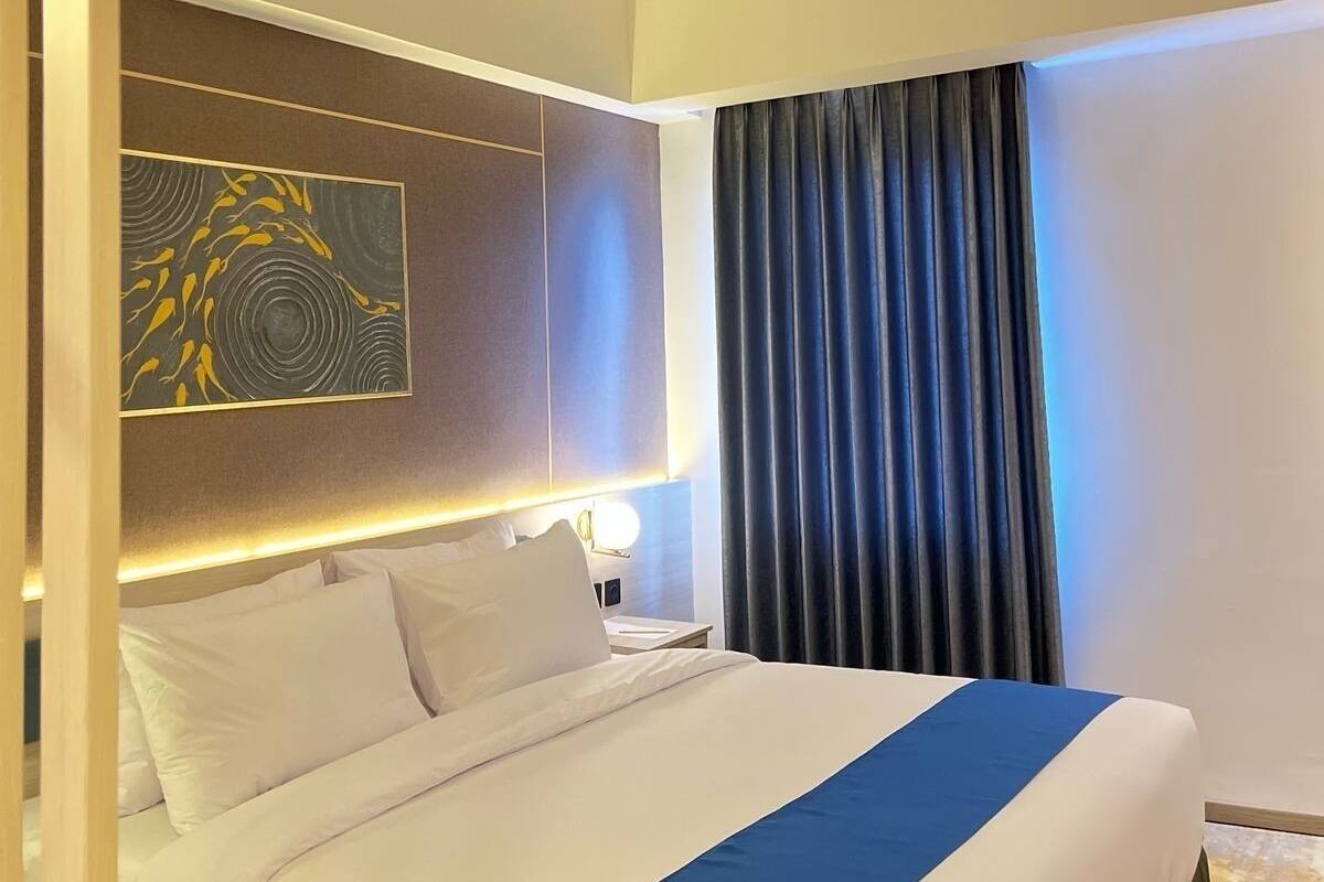 A hotel room with a large bed, a headboard with a light panel, a wall-mounted artwork, and a window with curtains.