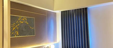 A hotel room with a large bed, a headboard with a light panel, a wall-mounted artwork, and a window with curtains.