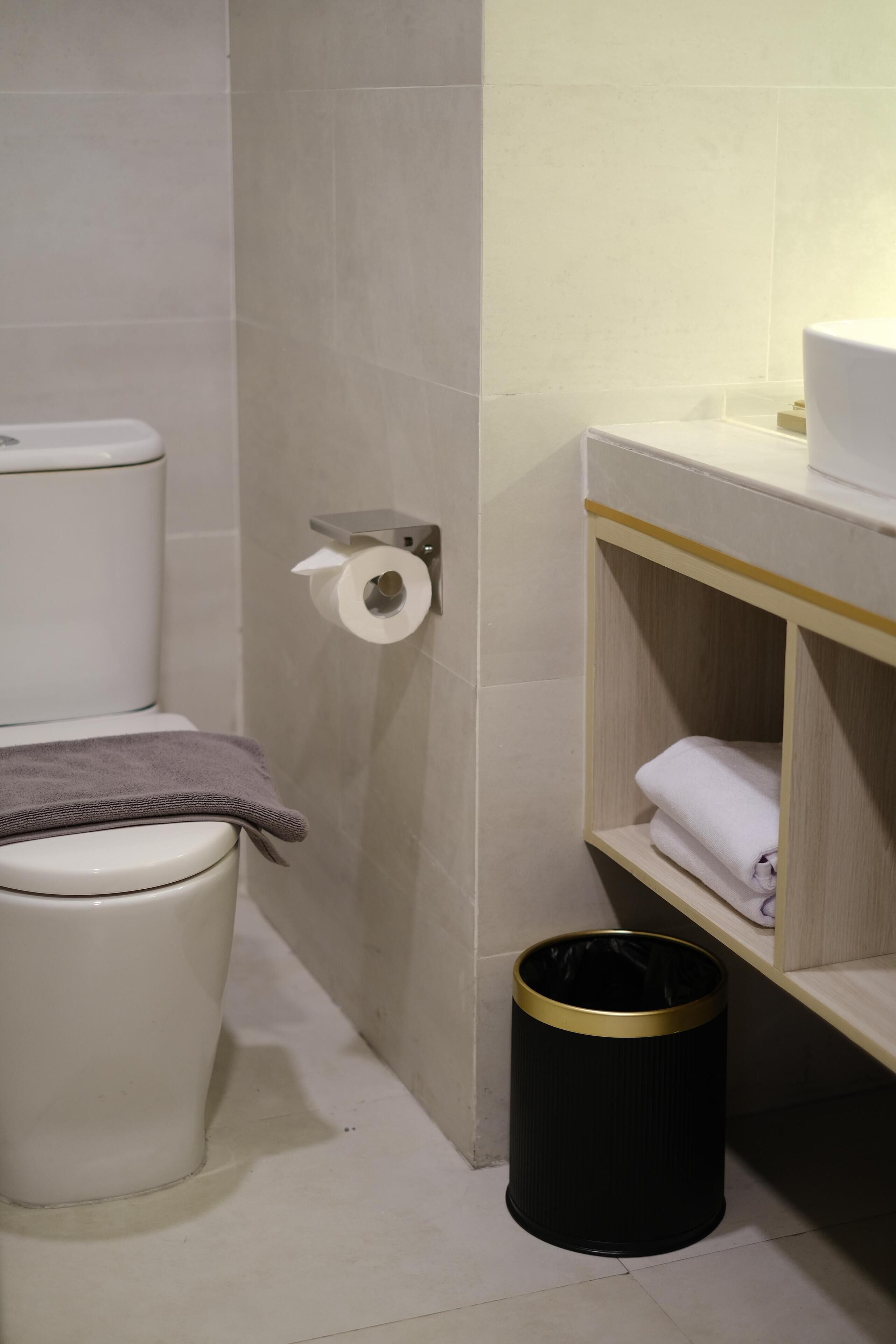 Un baño con inodoro, un rollo de papel higiénico en la pared, un cesto de basura negro y un estante con toallas dobladas.