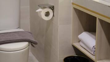 Un baño con inodoro, un rollo de papel higiénico en la pared, un cesto de basura negro y un estante con toallas dobladas.