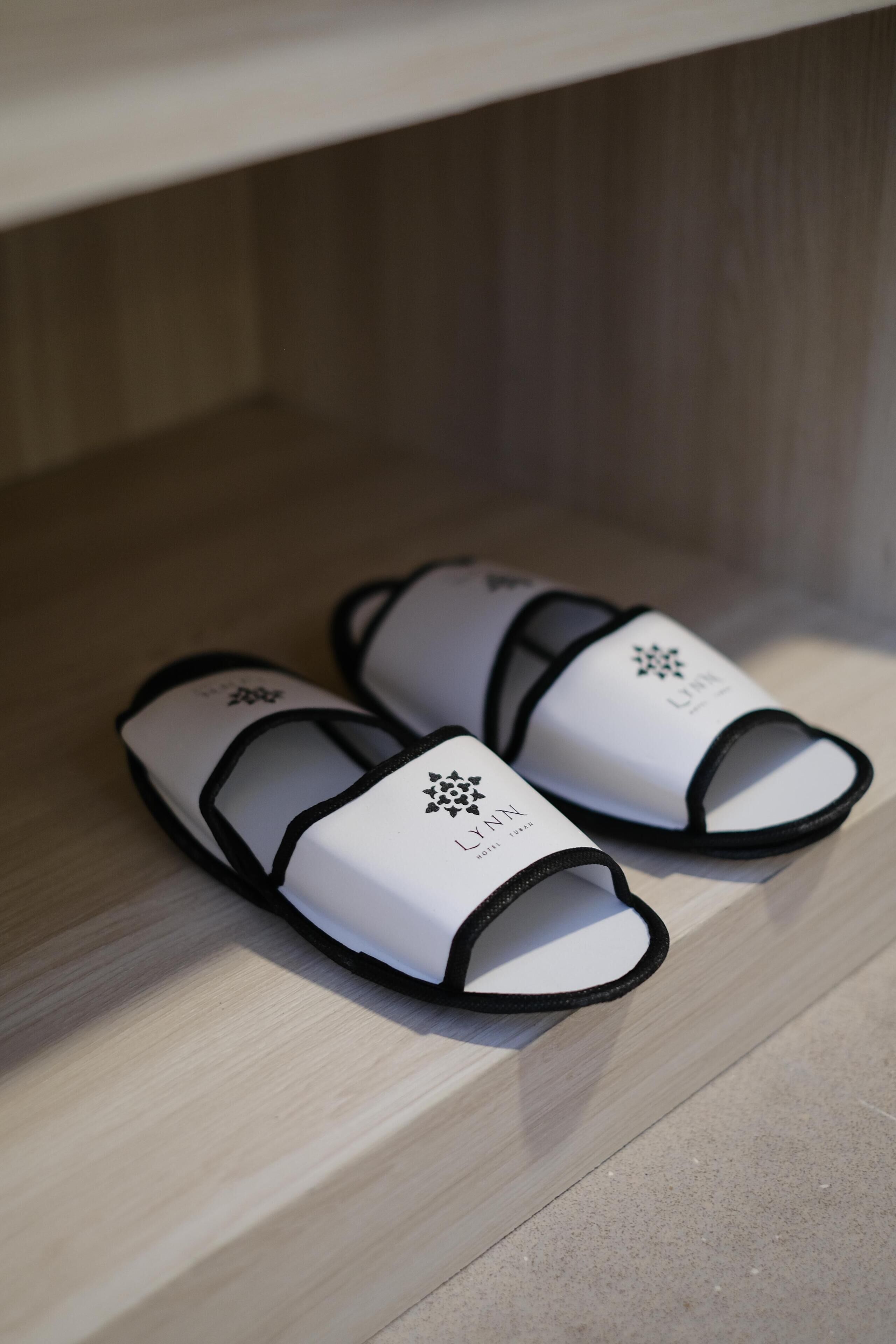 To hvide og sorte slippers med et logo foran, stående på et trægulv.