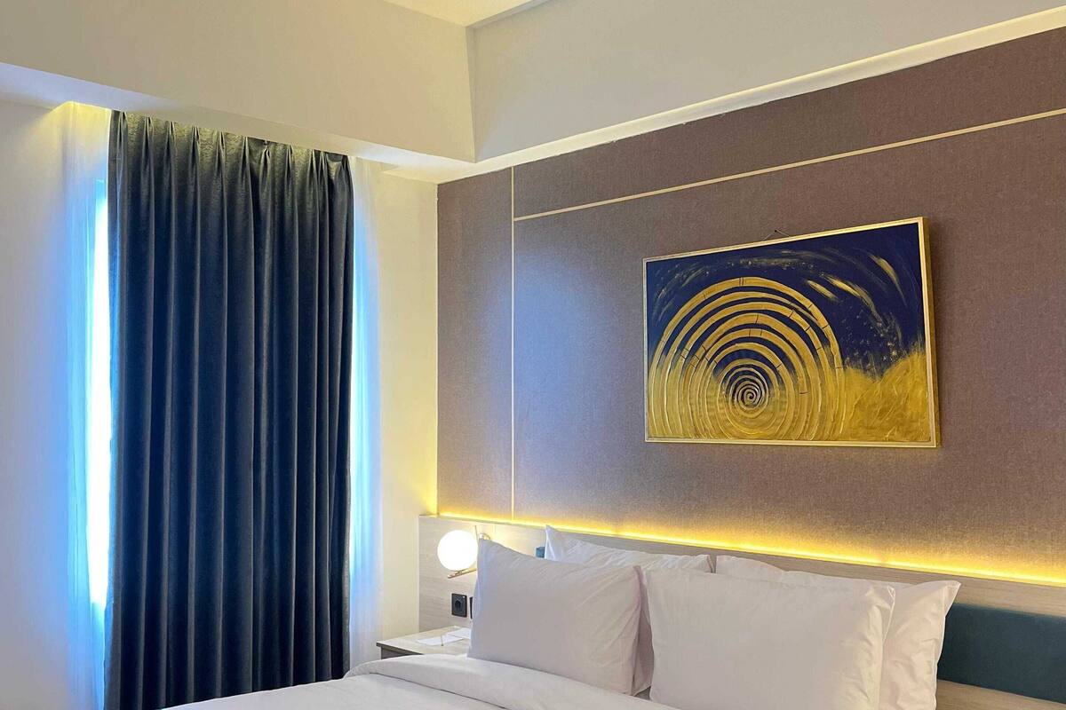 A hotel room with a large bed, a nightstand with a lamp, a painting on the wall, and a window with curtains.