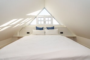 A bedroom with a sloped ceiling, a window with white shutters, and a bed with white bedding and two pillows.