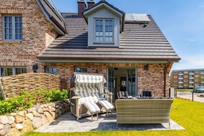Una casa de ladrillo con techo inclinado, un porche cubierto con mobiliario exterior y un muro de piedra.