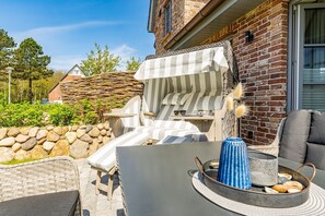Un espace extérieur aménagé avec des sièges, un mur en pierre, une chaise en osier, un parasol rayé et une table sur laquelle se trouvent un vase bleu et un panier de pain.