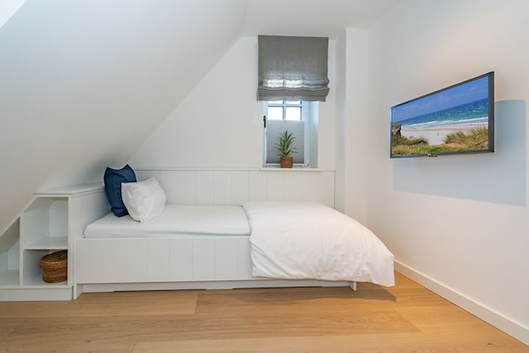 Une chambre avec un plafond en pente, un lit blanc, une petite fenêtre avec une plante et un tableau représentant une scène de plage au mur.