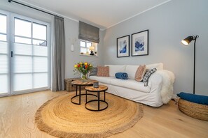 Un salon moderne avec un canapé blanc, deux petites tables rondes en bois, un tapis tissé et une grande fenêtre avec des stores.