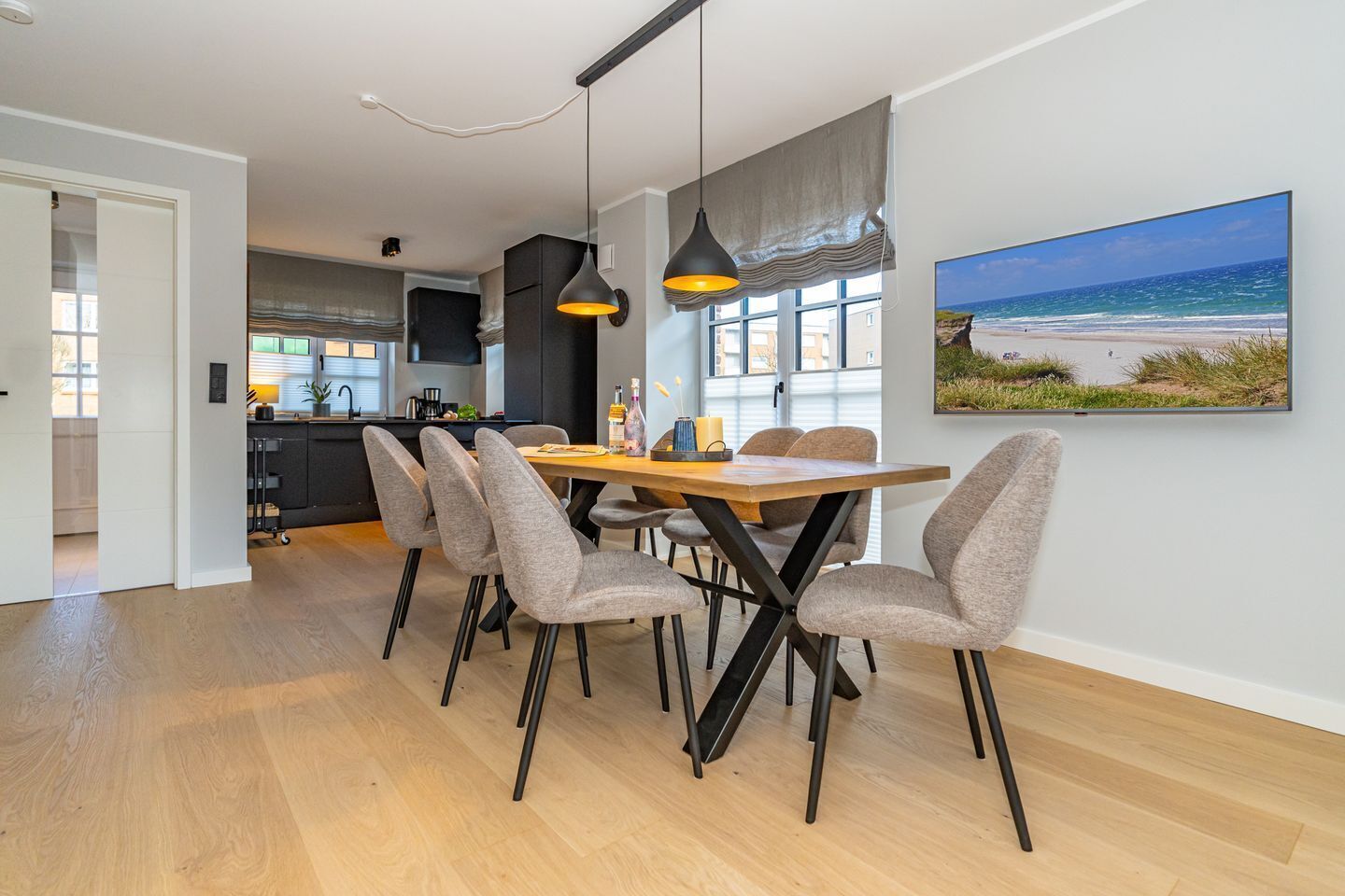 A modern dining area with a wooden table and grey upholstered chairs, a pendant light, and a large window with a beach view.
