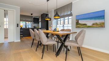 A modern dining area with a wooden table and grey upholstered chairs, a pendant light, and a large window with a beach view.