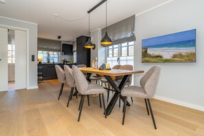 Un espace repas moderne avec une table en bois et des chaises à revêtement gris, un luminaire suspendu et une grande fenêtre donnant sur la plage.