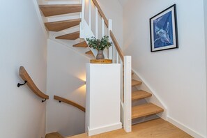 Un escalier avec des marches en bois et des murs blancs, une plante sur le palier et une œuvre d’art encadrée au mur.