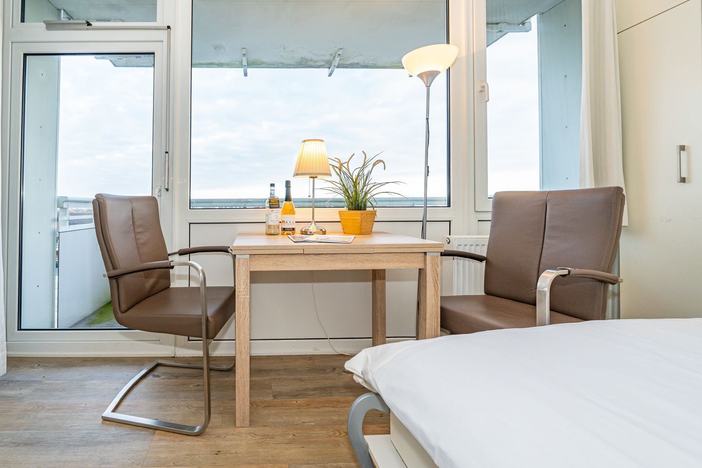Habitación de hotel con cama, dos sillas, una mesita con lámpara, una planta en maceta y vistas al mar a través de amplias ventanas.