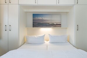 Quarto com cama, duas almofadas, pintura de uma praia e armários brancos.