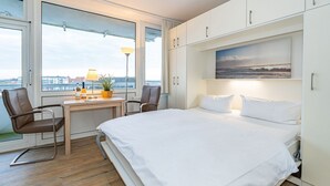 Moderni hotellihuone, jossa on suuri sänky, työpöytä tuolilla, ikkuna, josta avautuu näkymä kaupunkiin, ja taulu seinällä.