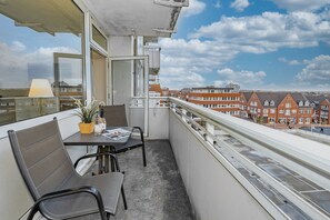 Un balcon avec une table et des chaises, une plante en pot et une bouteille. Le balcon donne sur une rue bordée d’immeubles en briques rouges.