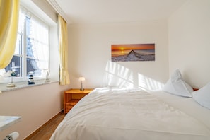Een slaapkamer met een bed, nachtkastje, lamp, raam met gordijnen en een schilderij van een zonsondergang.