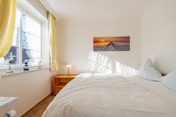 Une chambre à coucher comprenant un lit, une table de chevet, une lampe, une fenêtre avec des rideaux et un tableau représentant un coucher de soleil.