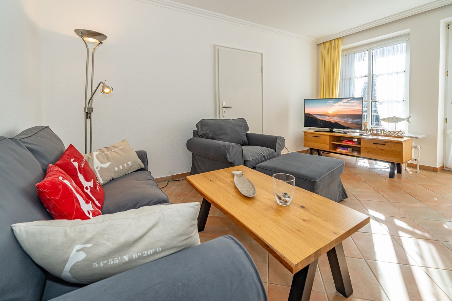 Ein modernes Wohnzimmer mit einem grauen Sofagarnitur, einem Holztisch und einem Fernseher auf einem Holzgestell.