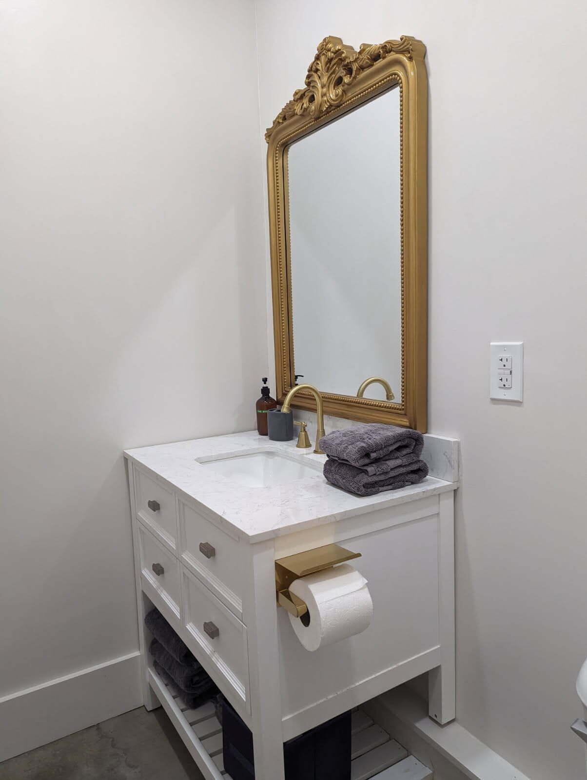 A bathroom with a white vanity, a gold-framed mirror, a sink, and a toilet.