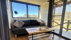 A modern living room with a wooden coffee table, a dark grey sofa with cushions, and a view of the outdoors through large windows.