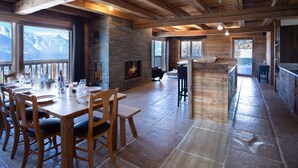 Une salle à manger spacieuse avec une cheminée en pierre, des poutres en bois et de grandes fenêtres donnant sur un paysage montagneux.