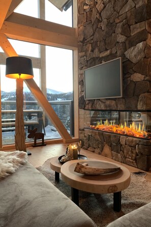 A cozy living room with a stone fireplace, a television mounted on the wall, and a large window overlooking a snowy landscape.
