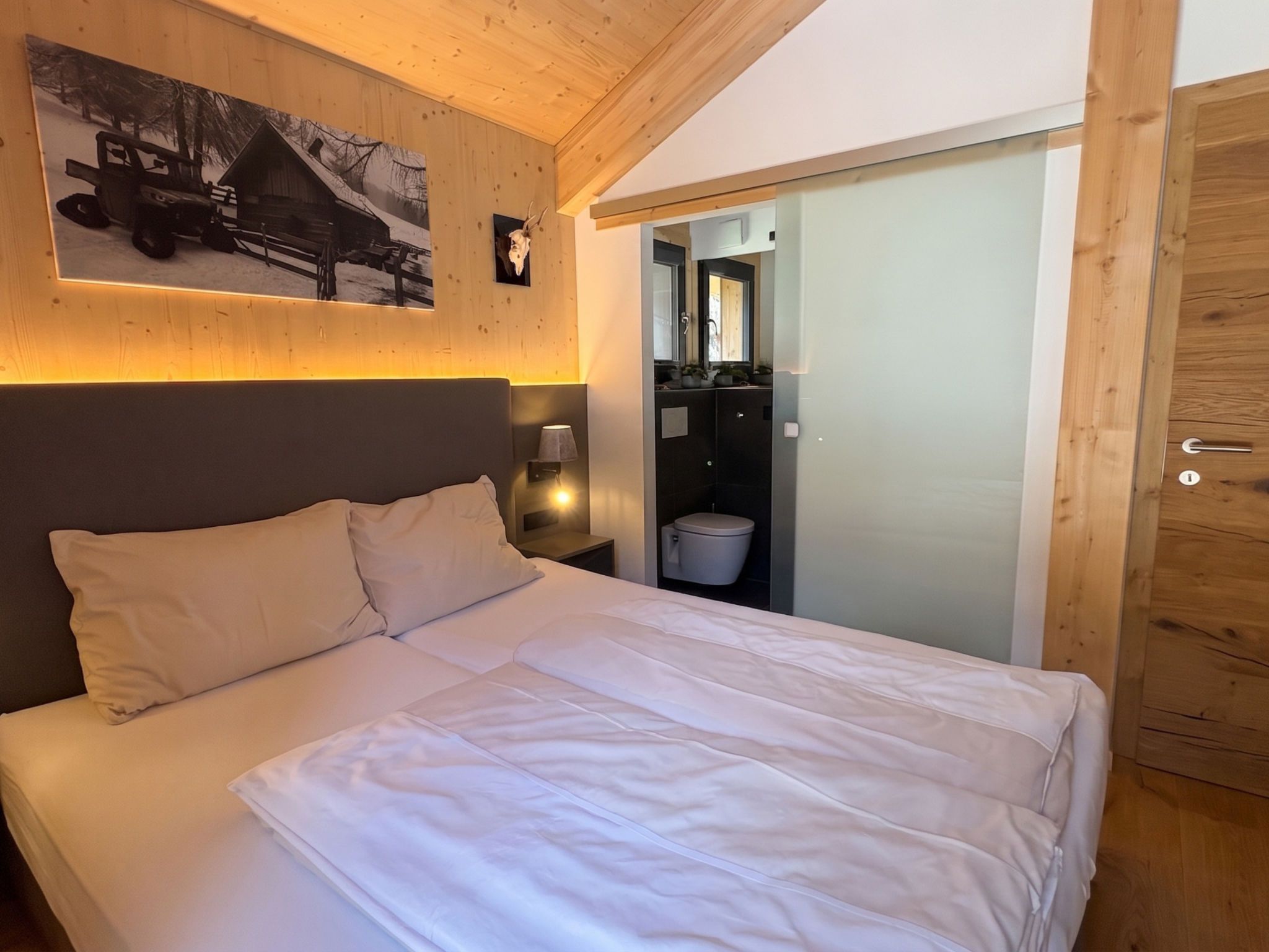 A bedroom with a bed, a nightstand, a wall-mounted lamp, a wooden door, and a bathroom visible through an open door.