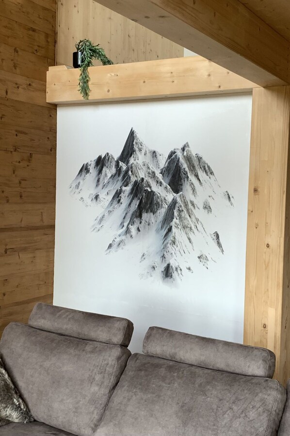 A room with a large painting of a mountain landscape on the wall, a wooden ceiling, and a grey sofa.