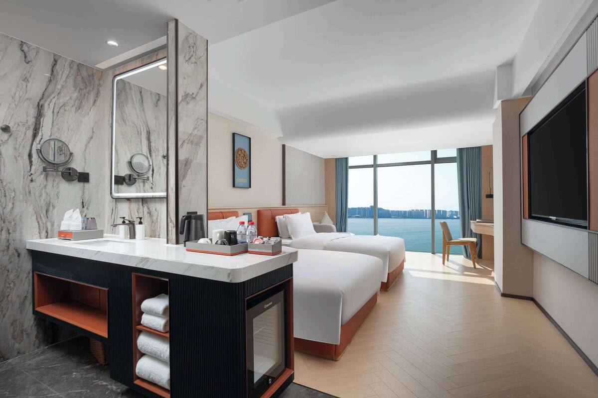 Ein Hotelzimmer mit Marmorbad, einem großen Spiegel, einem Bett, einem Fernseher und Blick aufs Wasser.
