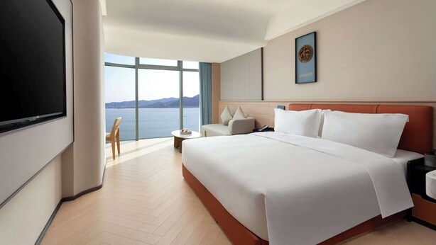 Ein Hotelzimmer mit einem großen Bett, einem an der Wand befestigten Fernseher, einem kleinen Tisch und Meerblick.