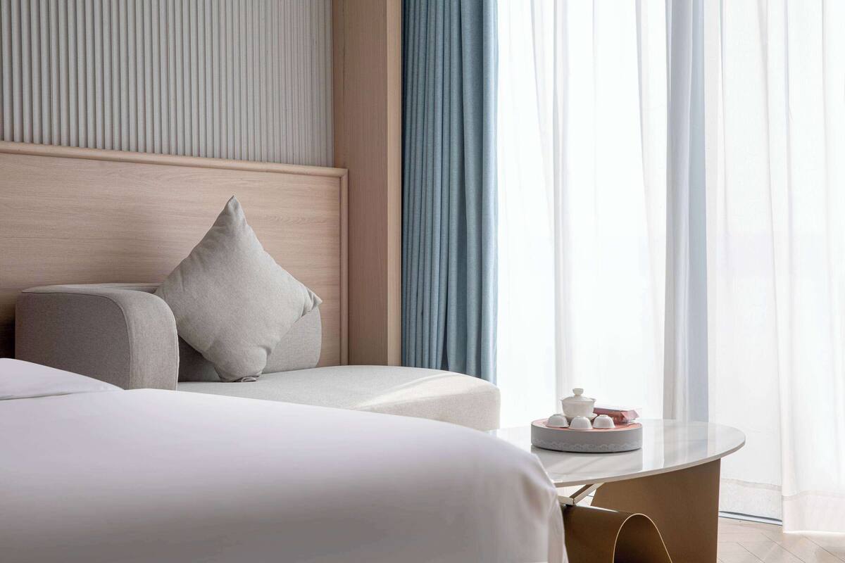 Ein modernes Hotelzimmer mit einem großen Bett, einem Nachttisch mit Dekoration und Blick ins Freie durch transparente Vorhänge.