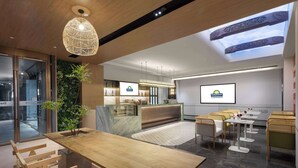 Eine moderne Hotelhalle mit Holzbude für die Rezeption, Sitzgelegenheiten und einem großen Bildschirm, der den Hotelnamen zeigt.