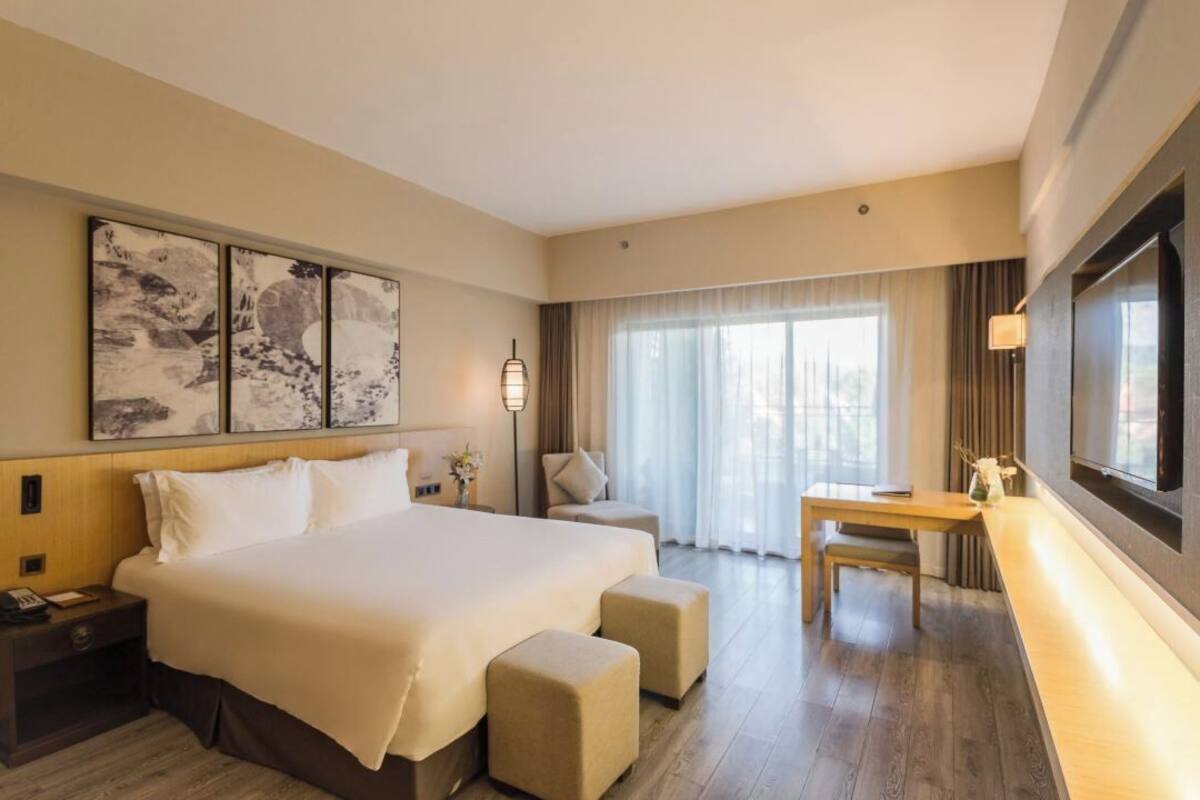 A hotel room with a large bed, a desk, and a chair. There is a window with curtains, a wall-mounted TV, and artwork above the bed.