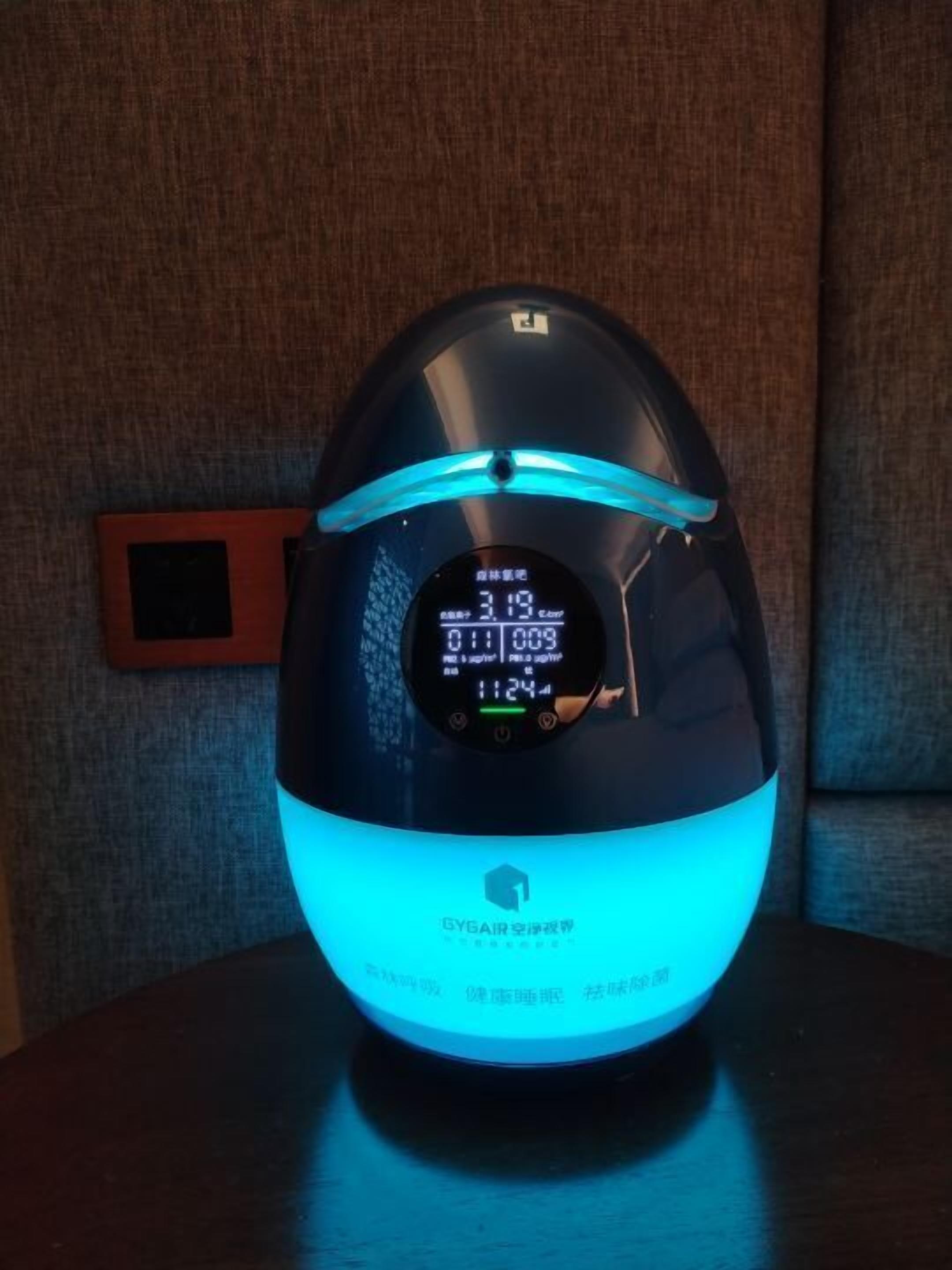 A smart speaker with a digital display, placed on a table against a textured wall.