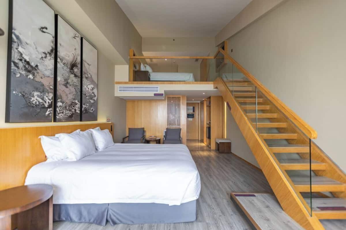 A modern hotel room with a large bed, a wooden staircase, and a view of the hallway.