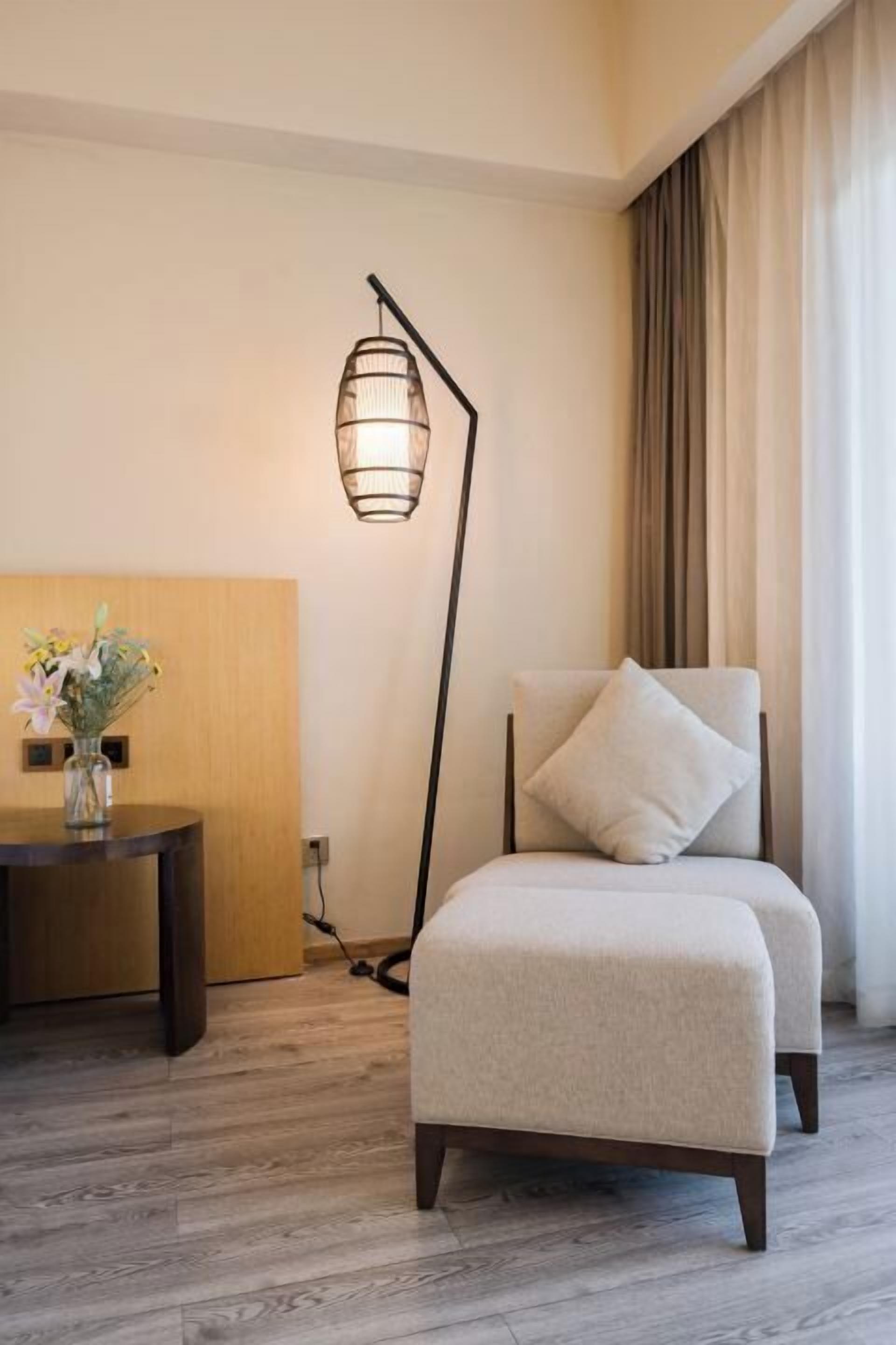 A modern hotel room with a wooden panel wall, a round table with a vase of flowers, a beige upholstered chair, and a floor lamp.