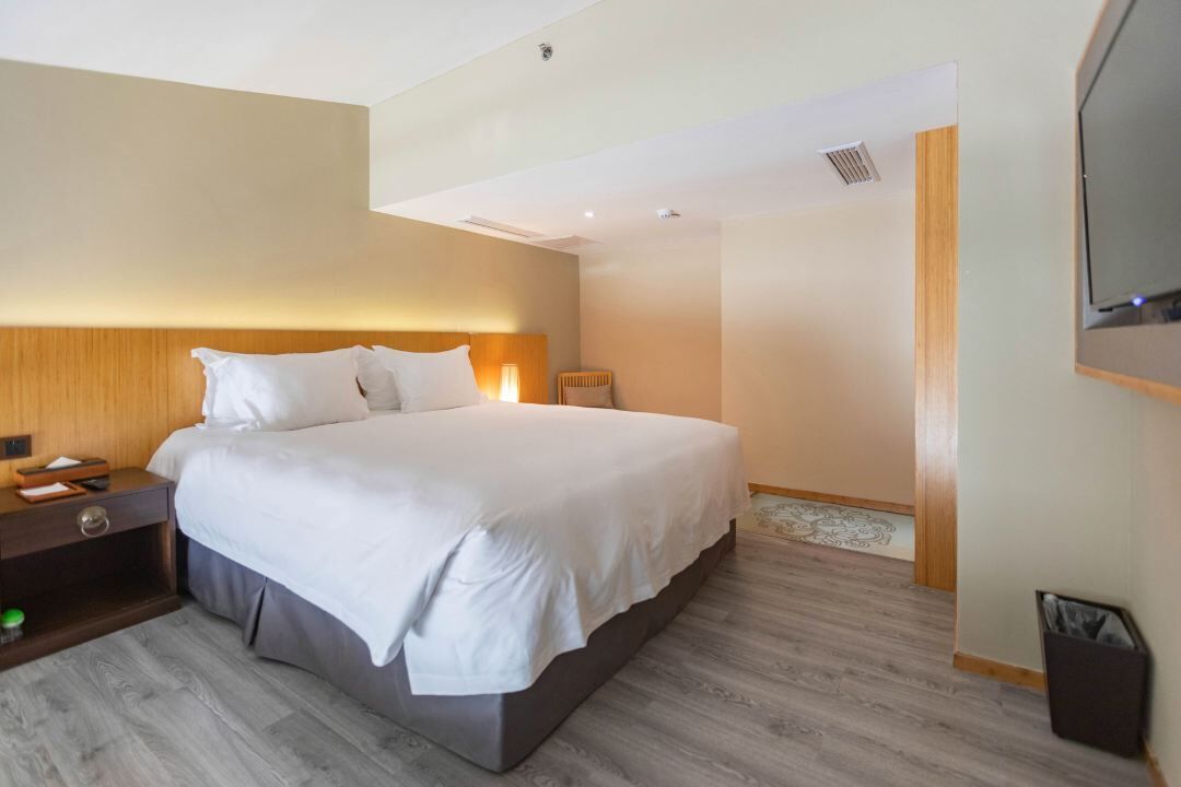A modern hotel room with a large bed, wooden headboard, bedside table, and a TV mounted on the wall.