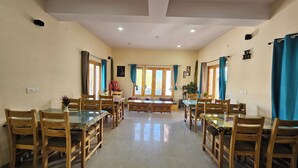 A dining area with wooden tables and chairs, a central bench, and large windows with curtains. - LUNGDHO RESIDENCY (Leh)