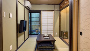A traditional Japanese room with tatami flooring, a low table with cushions, a TV, and a sliding door leading to a garden.