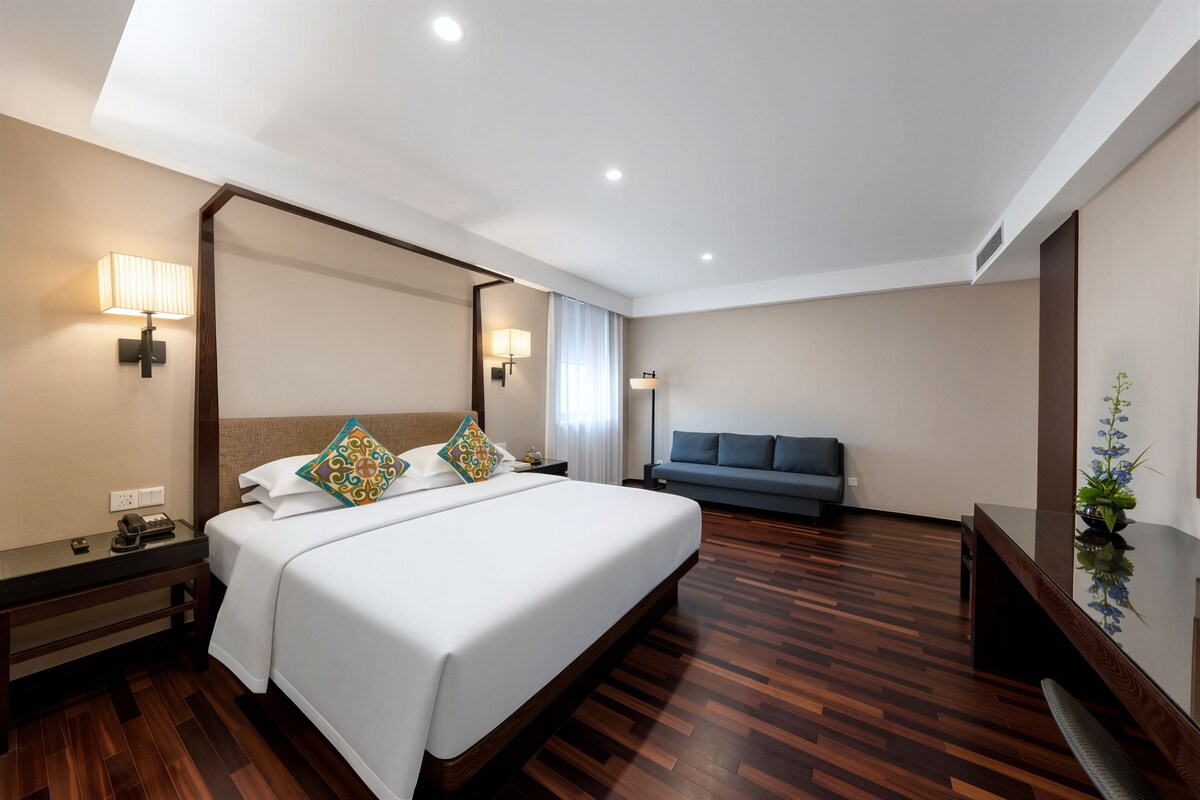 A hotel room with a bed, bedside table, lamp, and a sofa.