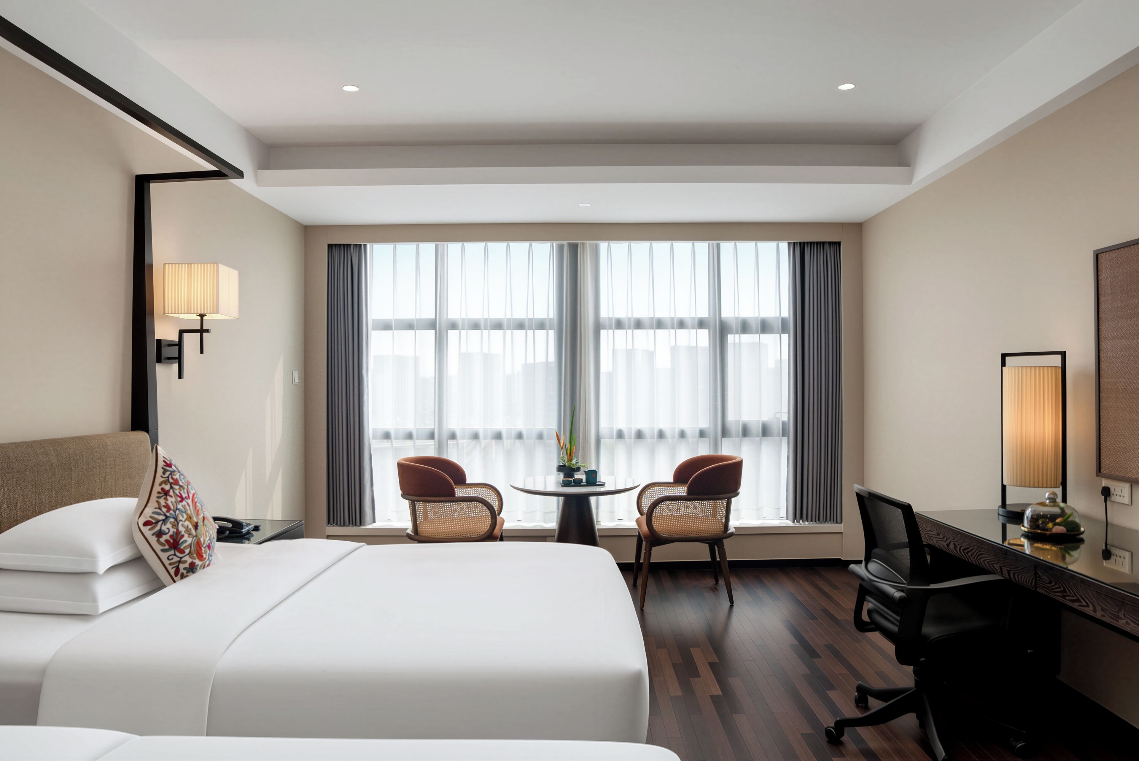 A hotel room with two beds, a desk, a chair, a lamp, and a view through large windows.