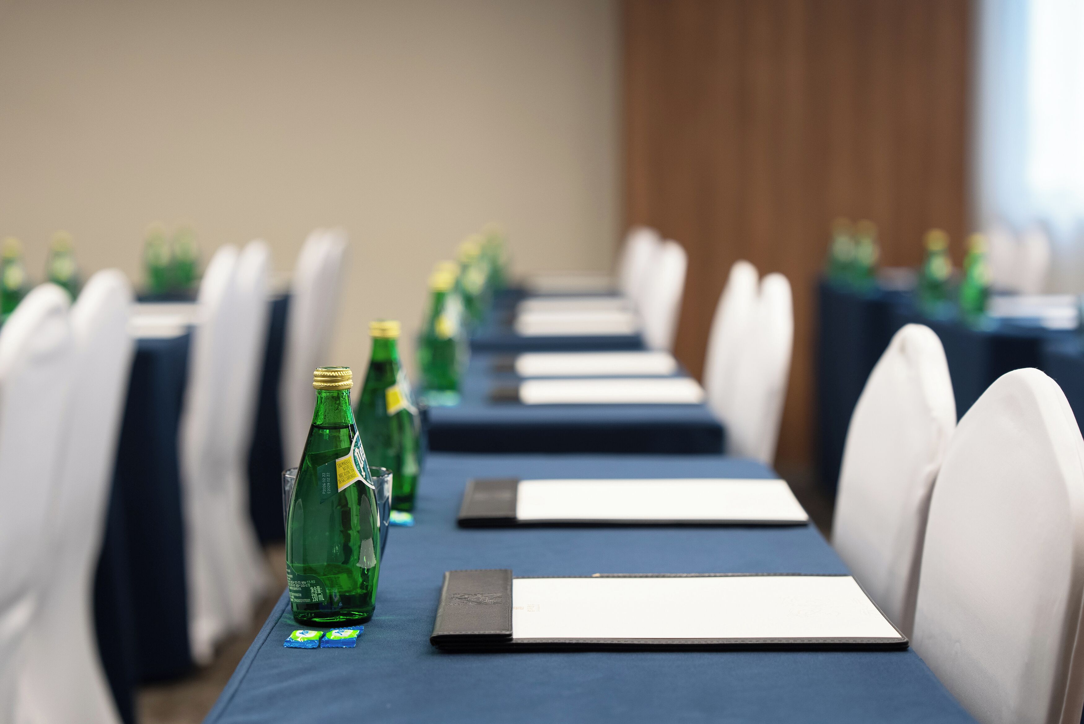 Un salón de conferencias con filas de sillas blancas, mesas con placas de identificación y botellas verdes sobre las mesas.