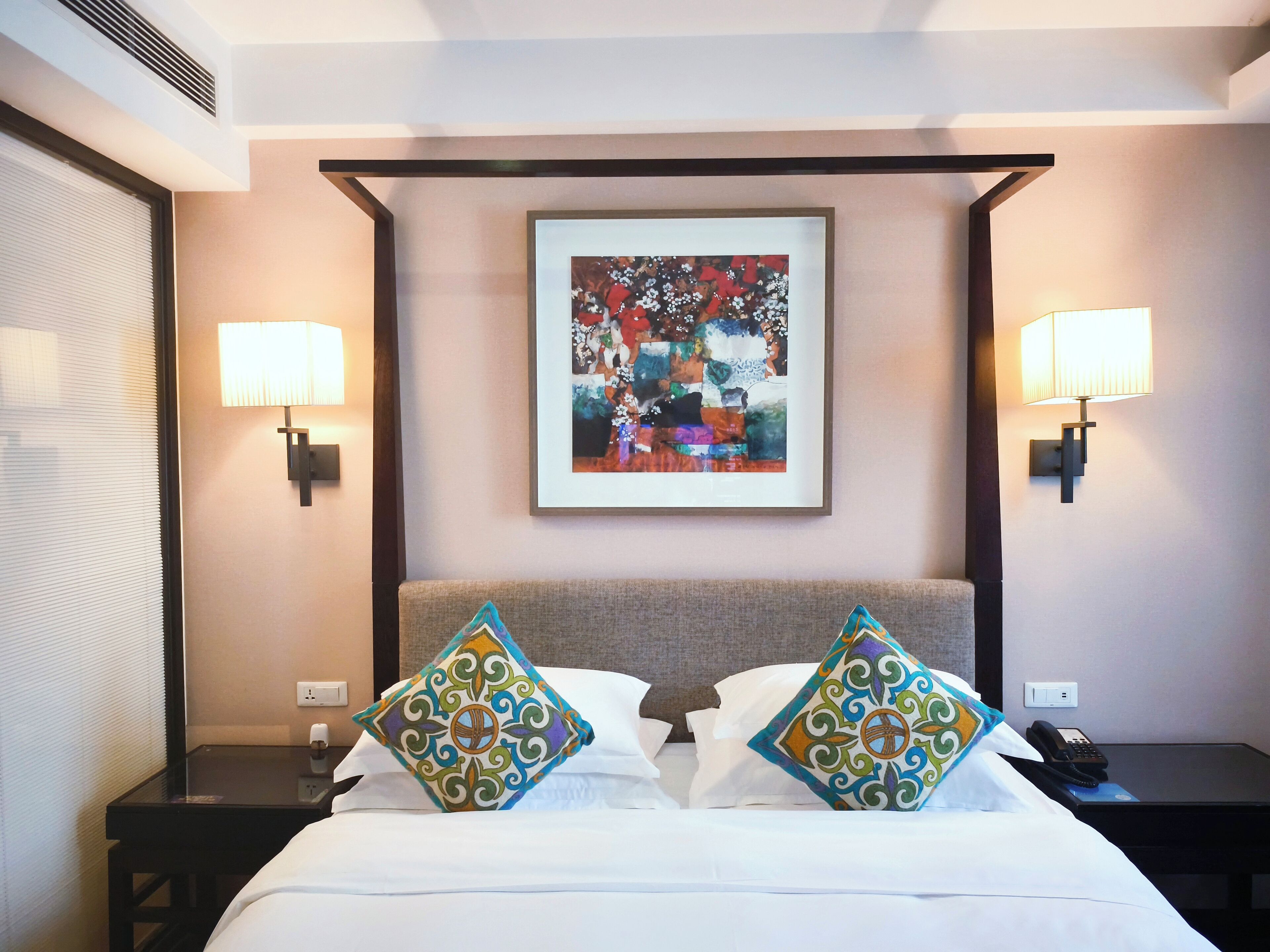 A hotel room with a bed, two bedside lamps, a framed abstract painting, and a nightstand with a phone.