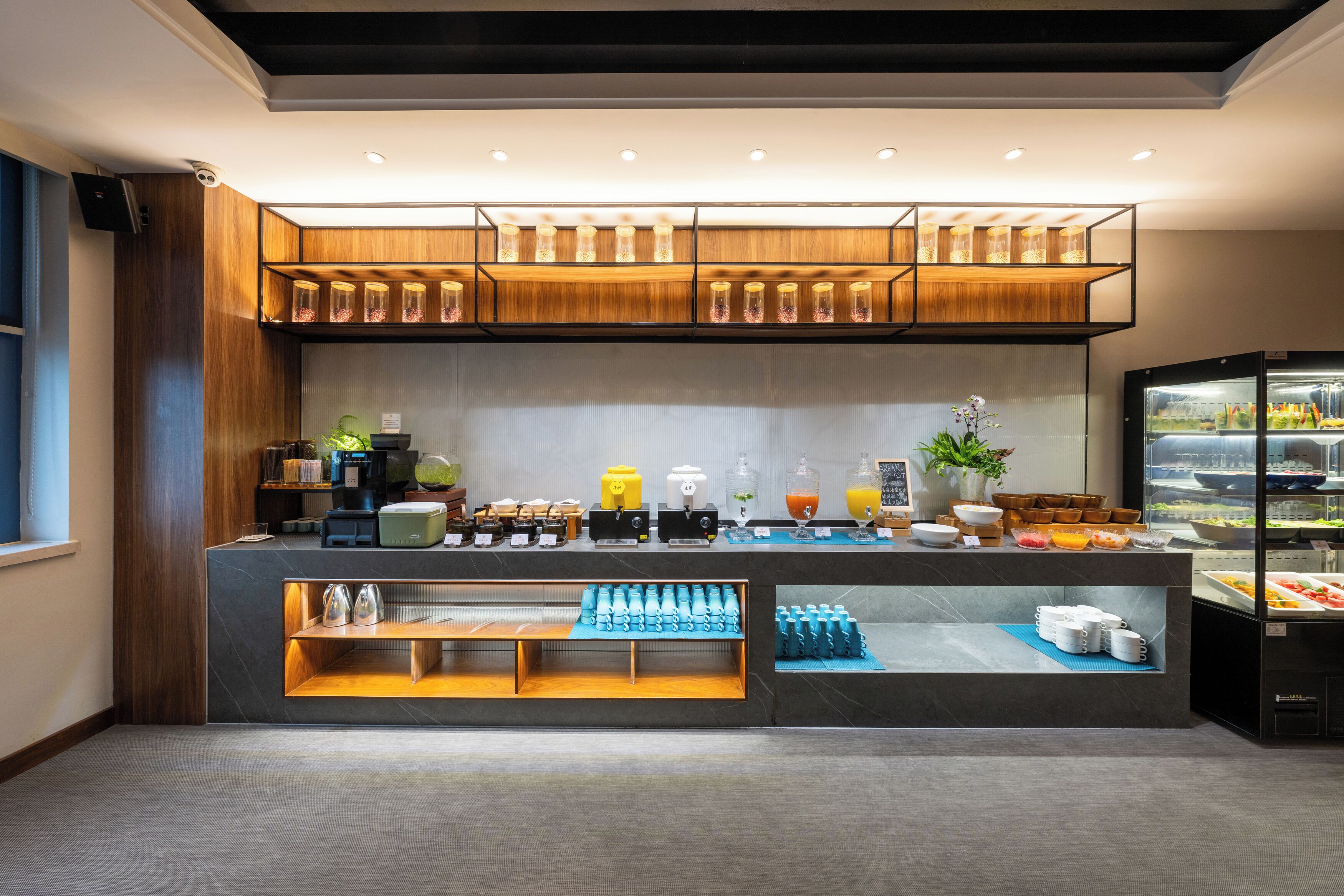 Una barra de desayuno moderna con diversas opciones de comida y bebida, una estación de café y un refrigerador con productos frescos.