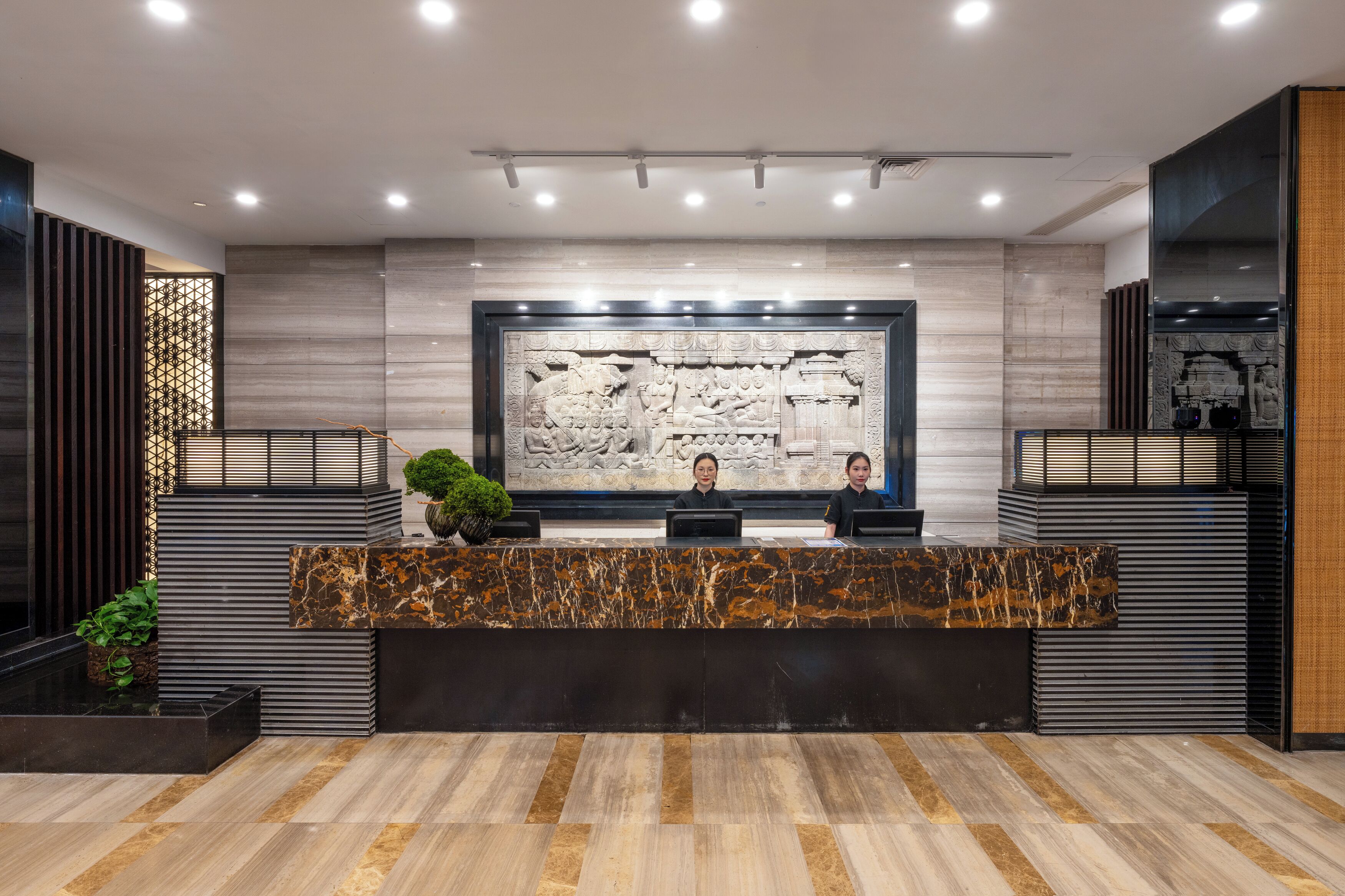 A reception desk with two staff members, a marble wall with a relief sculpture, and a modern interior design.