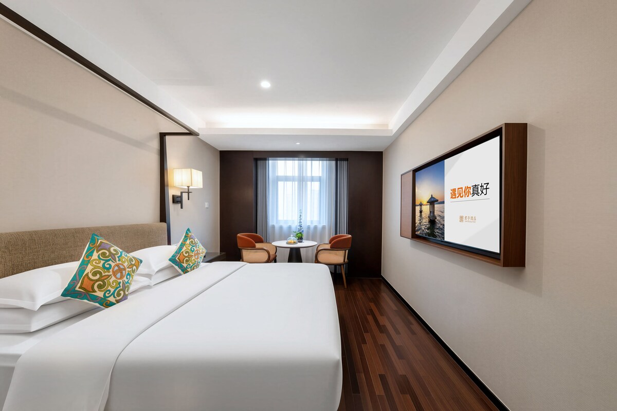 A hotel room with a large bed, two chairs, a small table, and a wall-mounted TV.