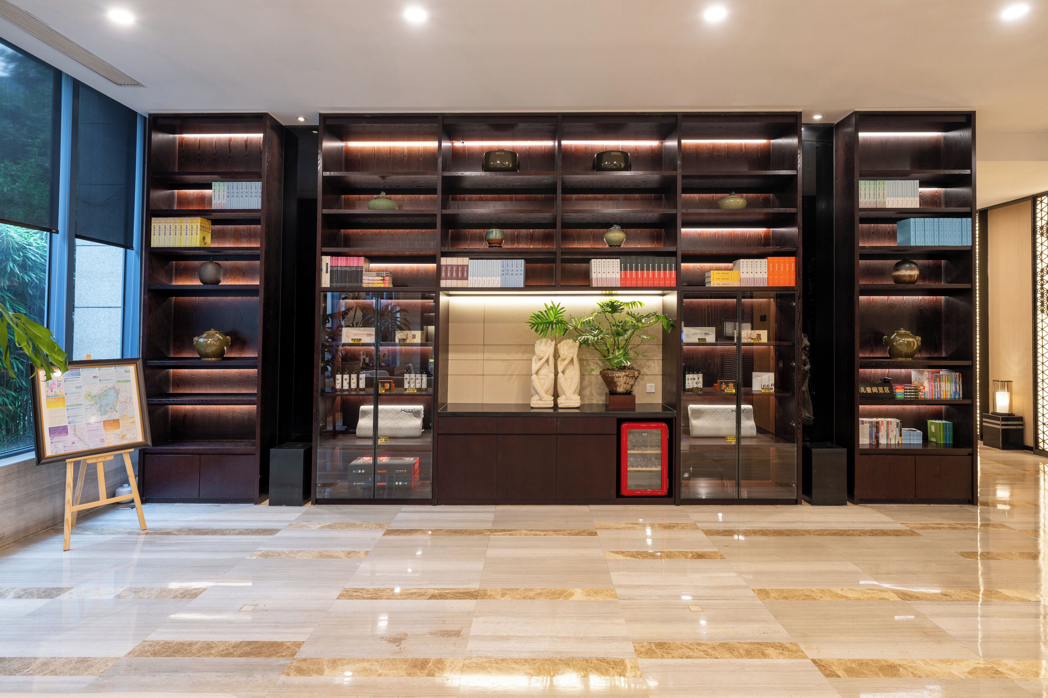 Un vestíbulo amplio con una gran estantería llena de libros y objetos decorativos, una pequeña zona de estar con una planta, y un mapa sobre un caballete.