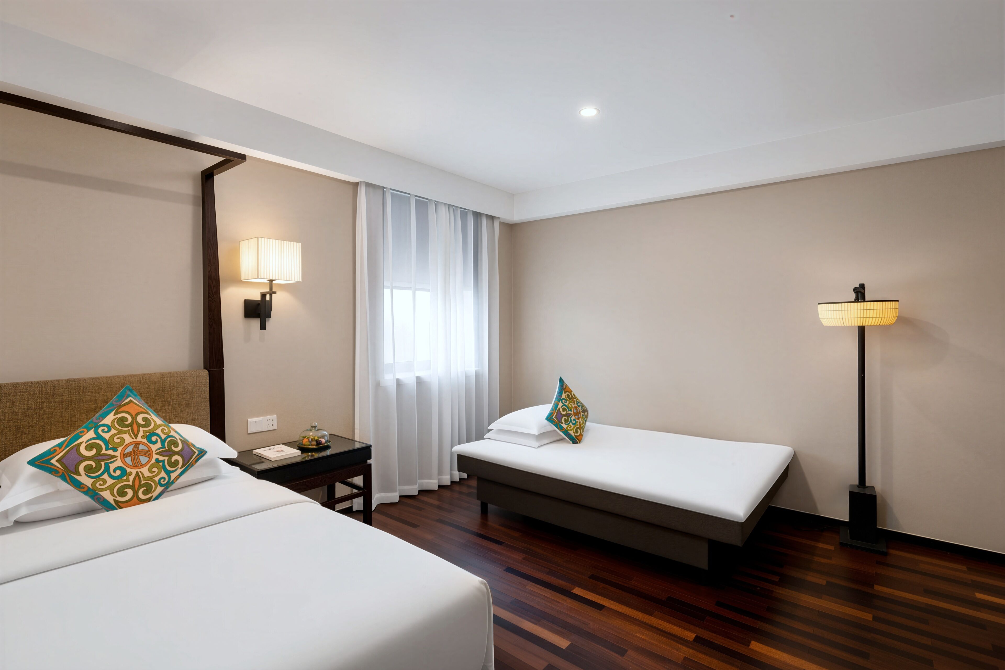 Quarto de hotel com duas camas, uma mesa de cabeceira, uma luminária e uma janela com cortinas.